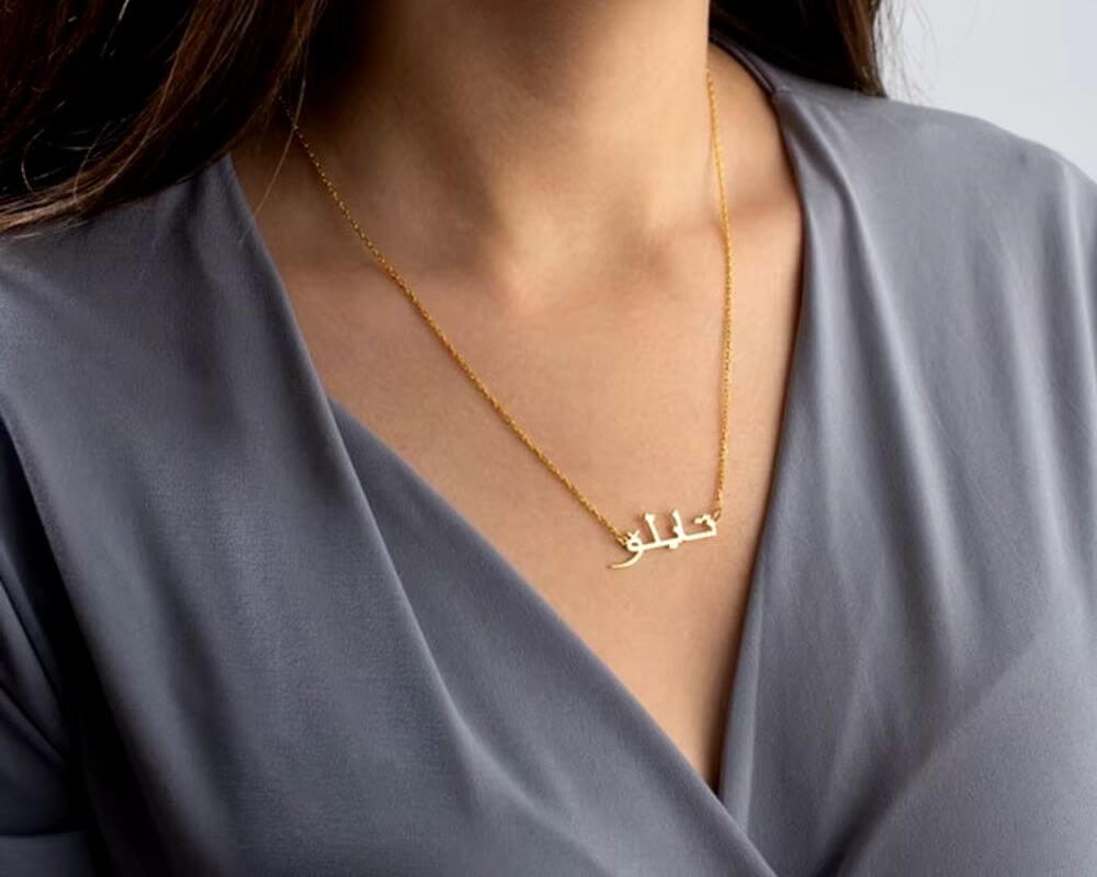 A gold necklace with a custom Arabic name design, elegantly styled against a minimalist gray blouse